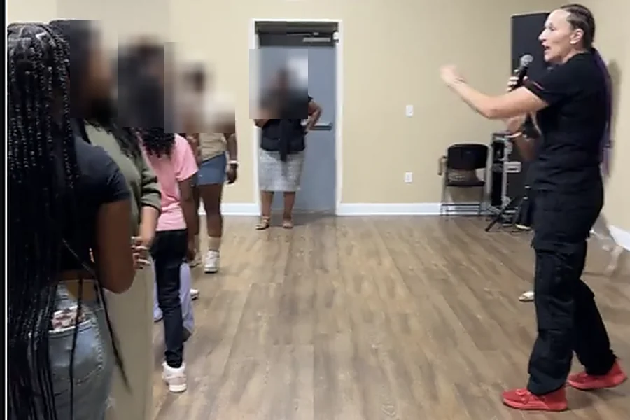 Carmen “The Momba” Gulliksen speaking with a microphone to a group at a community session; the attendees’ faces are blurred for their privacy